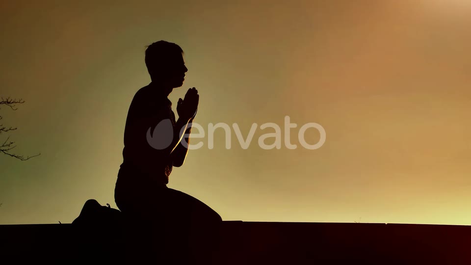 He kneels, hands folded, in silhouette against the sky
